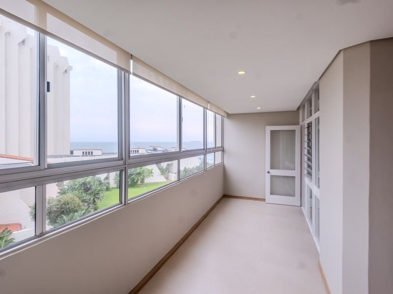 A bright, enclosed balcony featuring large windows with ocean views, tiled flooring, and recessed lighting, offering a versatile space for relaxation.
