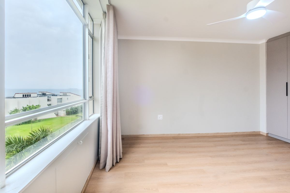 A bright, empty bedroom featuring light laminate flooring, a built-in wardrobe, a ceiling fan, and large windows offering a pleasant view.