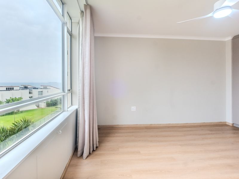 A bright, empty bedroom featuring light laminate flooring, a built-in wardrobe, a ceiling fan, and large windows offering a pleasant view.