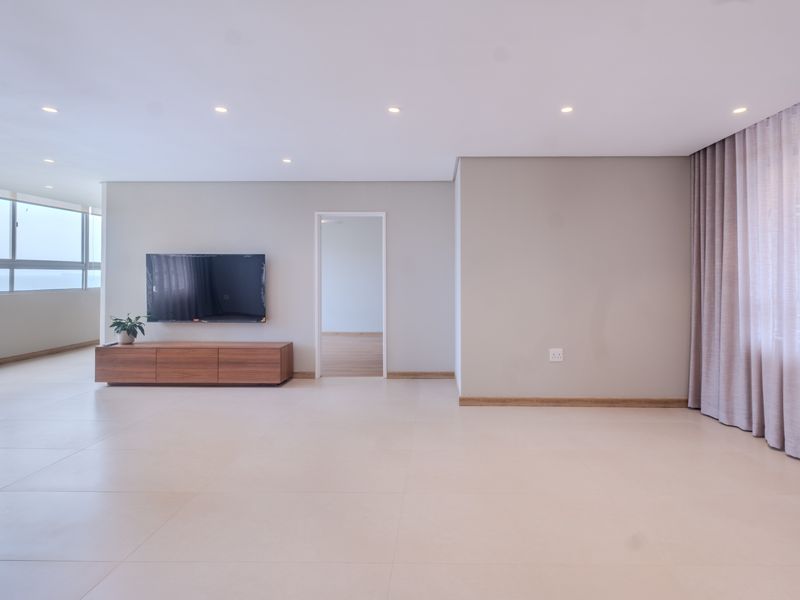 A spacious, light-filled living area featuring neutral large-format floor tiles, recessed downlighting, and elegant floor-to-ceiling sheer curtains.