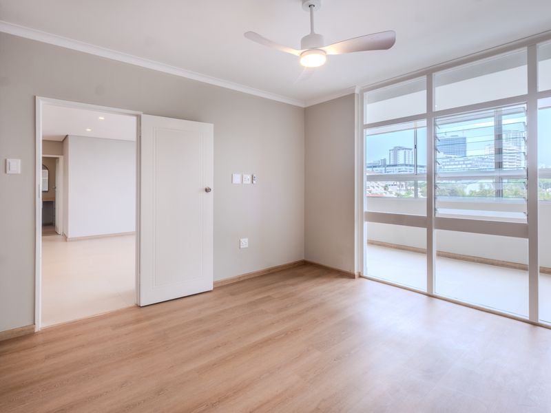 A bright, modern bedroom featuring light laminate flooring, a ceiling fan, built-in cupboards, and large glass doors leading to a balcony with city views.