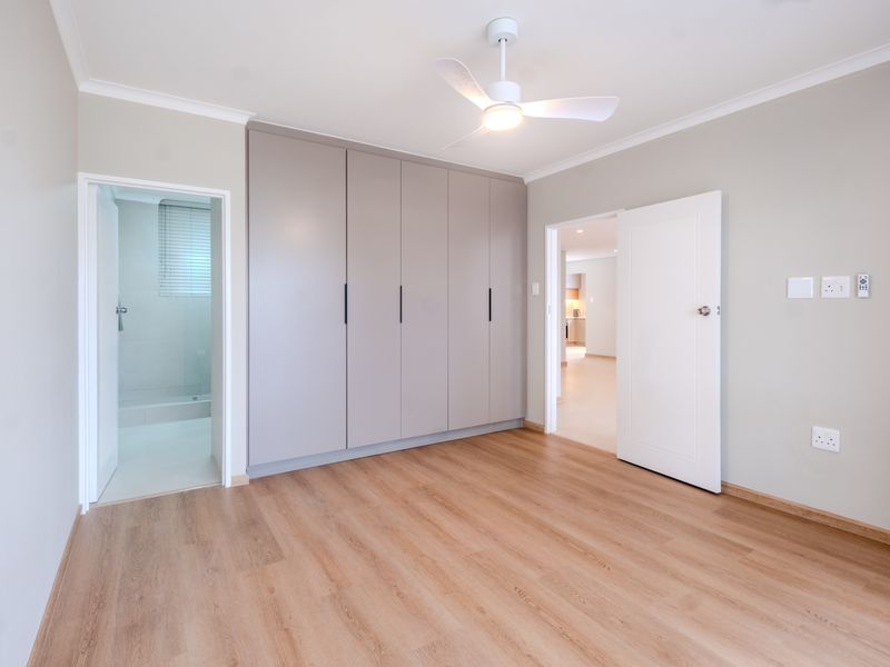 A bright, empty bedroom featuring modern laminate flooring, built-in cupboards, and a ceiling fan, with an adjoining en-suite bathroom.