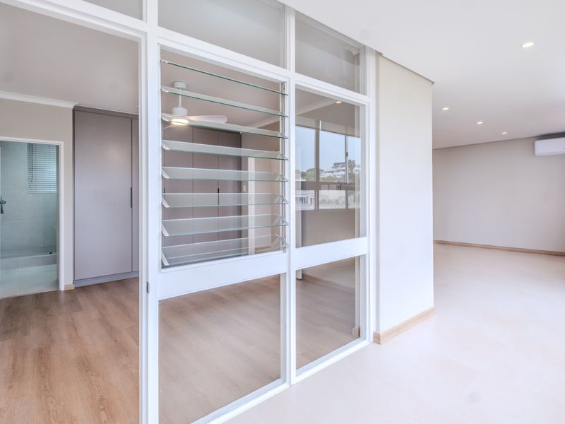 Bright open-plan living area featuring modern glass partitions, louvered windows, built-in storage, and neutral finishes throughout this well-maintained apartment space.