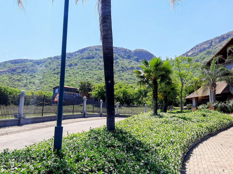 A well-maintained entrance area featuring a paved driveway, landscaped garden beds, and secure perimeter fencing with scenic mountain views in the background.