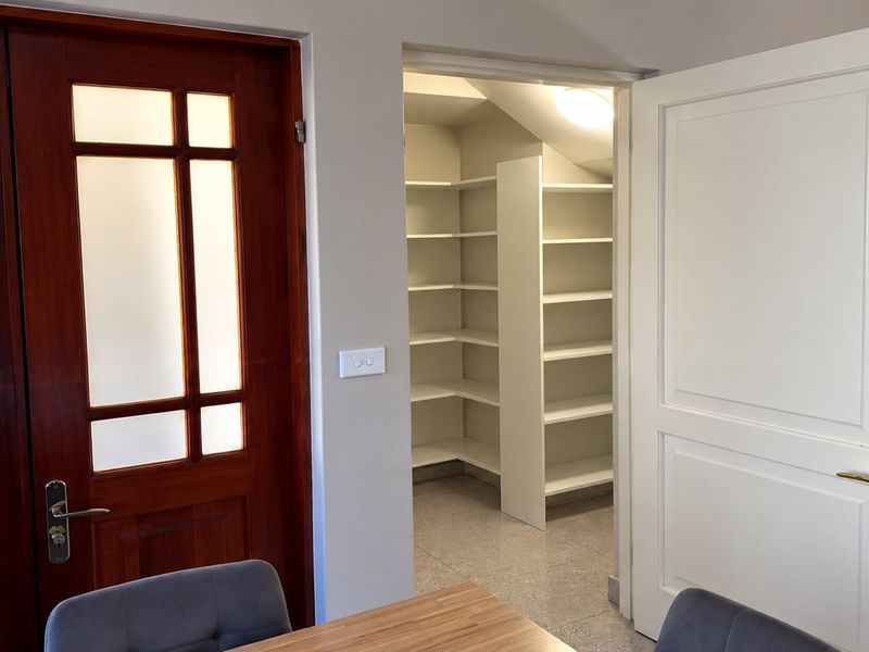 A clean, walk-in pantry or scullery area featuring extensive built-in shelving and tiled flooring, accessible via a white panel door.