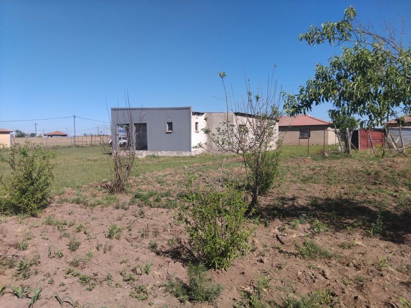 A view of the rear exterior of the house showing a mix of painted and unfinished masonry walls set within a large, undeveloped garden plot.