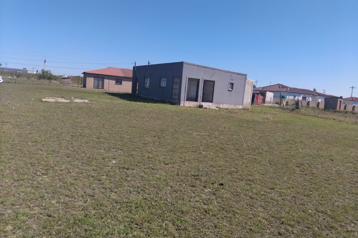 A detached single-story house viewed from the rear, featuring a large open yard, security gates, and metal window frames.