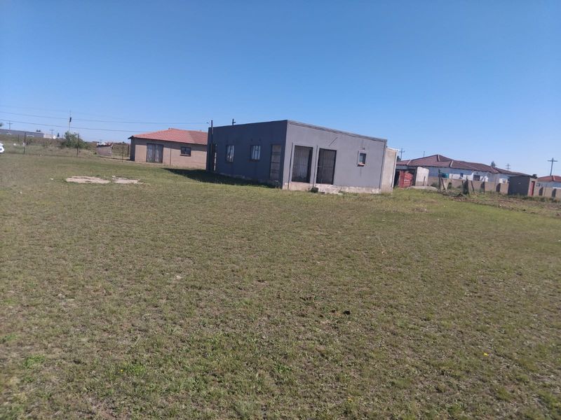 A detached single-story house viewed from the rear, featuring a large open yard, security gates, and metal window frames.