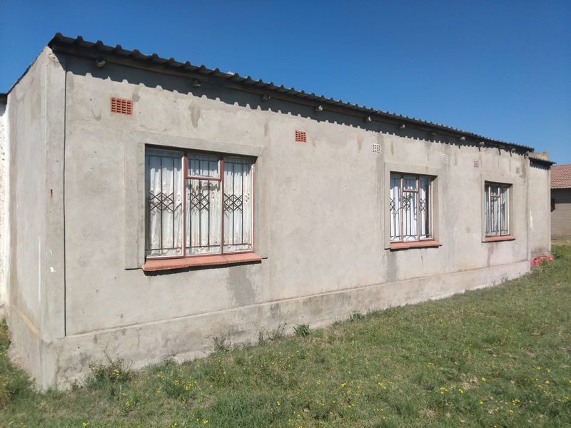 Rear exterior view of a single-story house featuring unpainted plastered walls, metal window frames with security bars, and a corrugated metal roof.