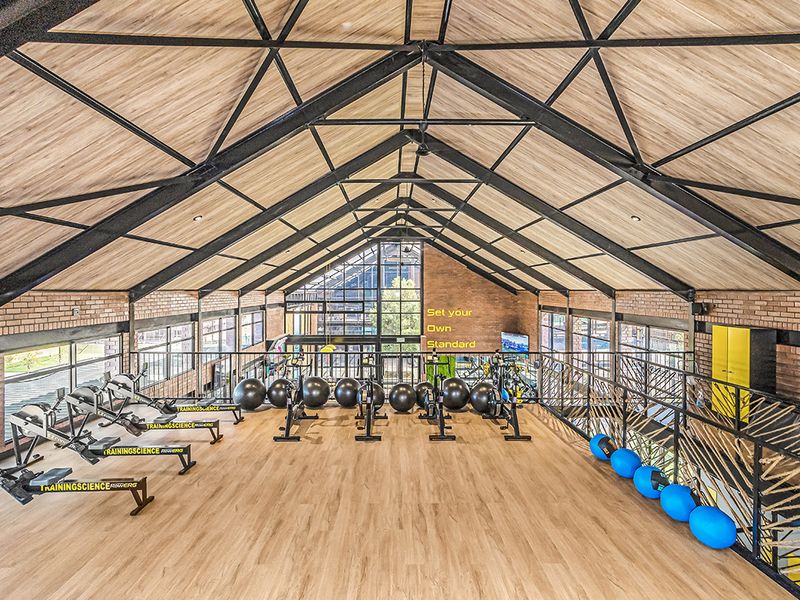 Spacious, modern gym featuring high vaulted ceilings with exposed steel trusses, brick feature walls, and large windows providing ample natural light.