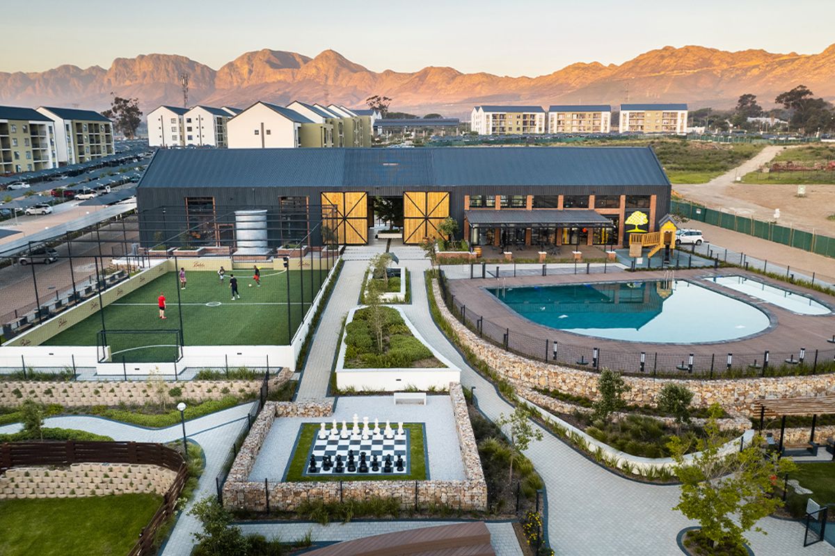 Aerial view of a modern apartment complex clubhouse featuring a swimming pool, sports court, landscaped gardens, and recreational facilities against a mountain backdrop.