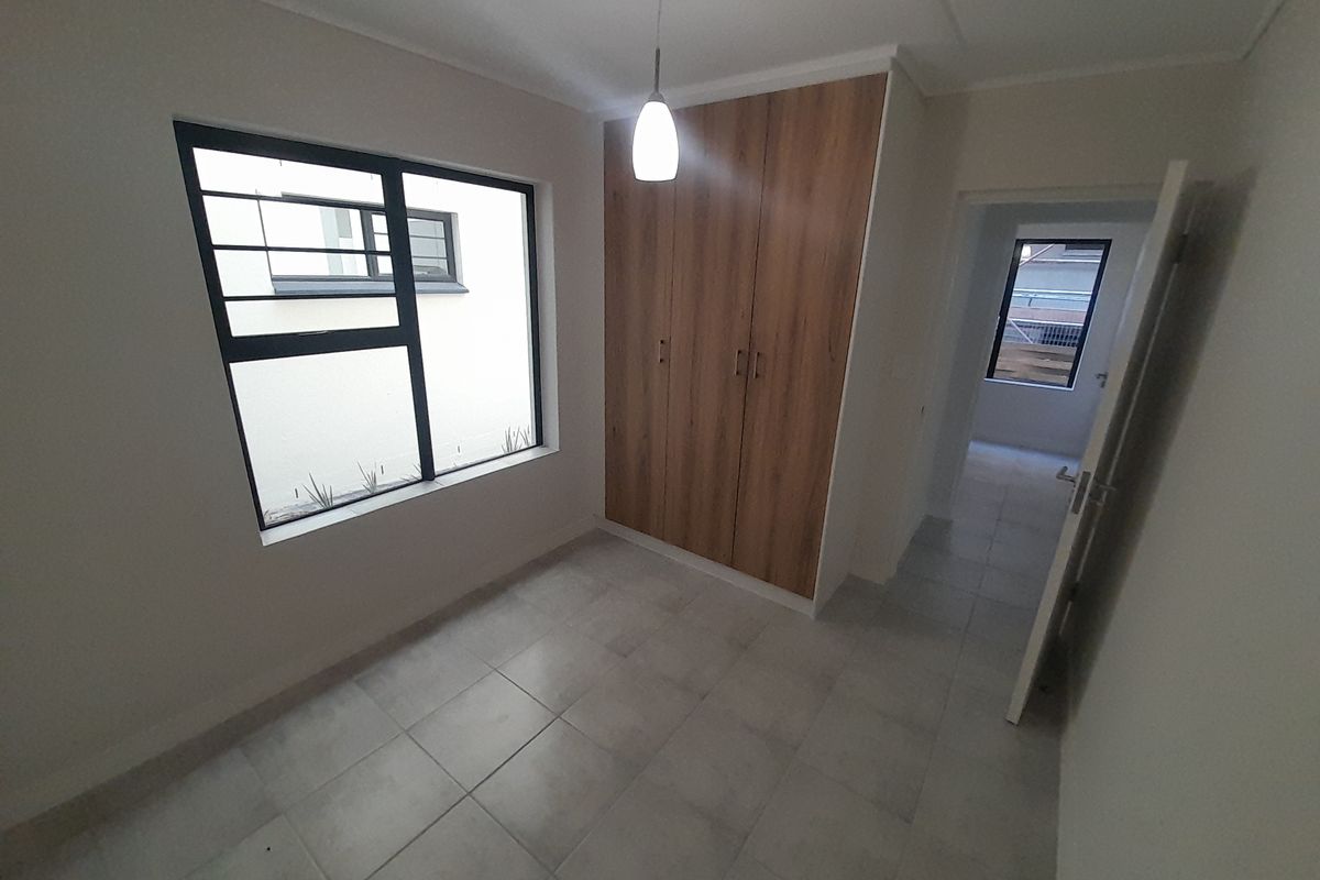 A clean, empty bedroom featuring modern built-in cupboards, neutral tiled flooring, and aluminium window frames with security bars.