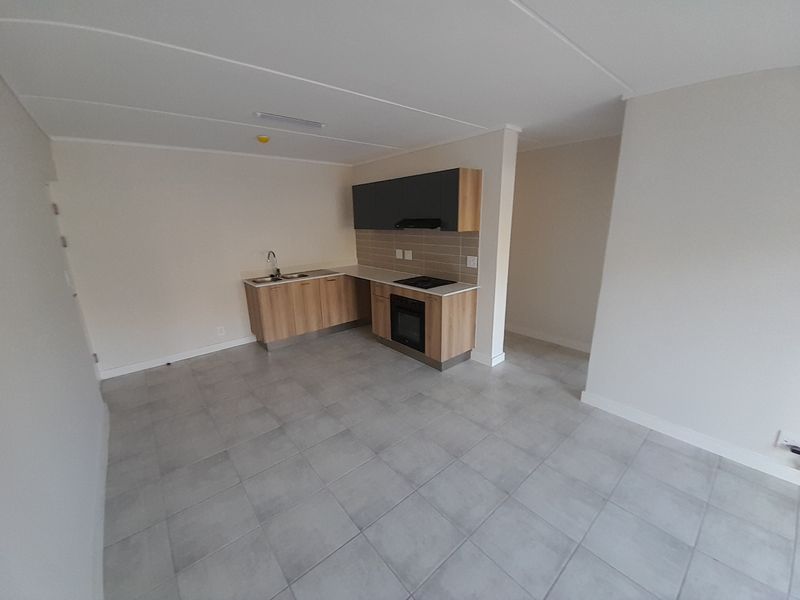 An open-plan living and kitchen area featuring modern light wood cabinetry, integrated appliances, and neutral tiled flooring in a clean, vacant space.