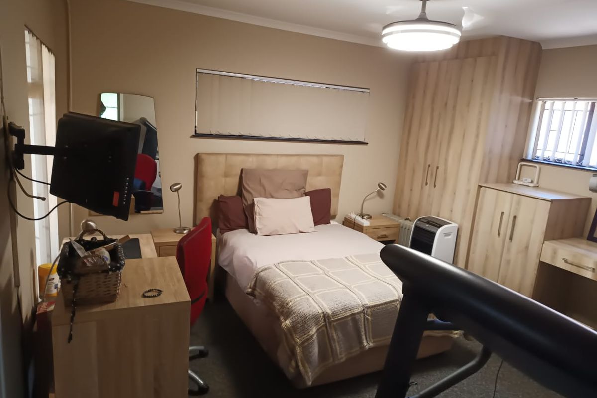 A functional bedroom featuring built-in wooden cupboards, a ceiling fan, and security bars on the window, providing a practical and secure living space.