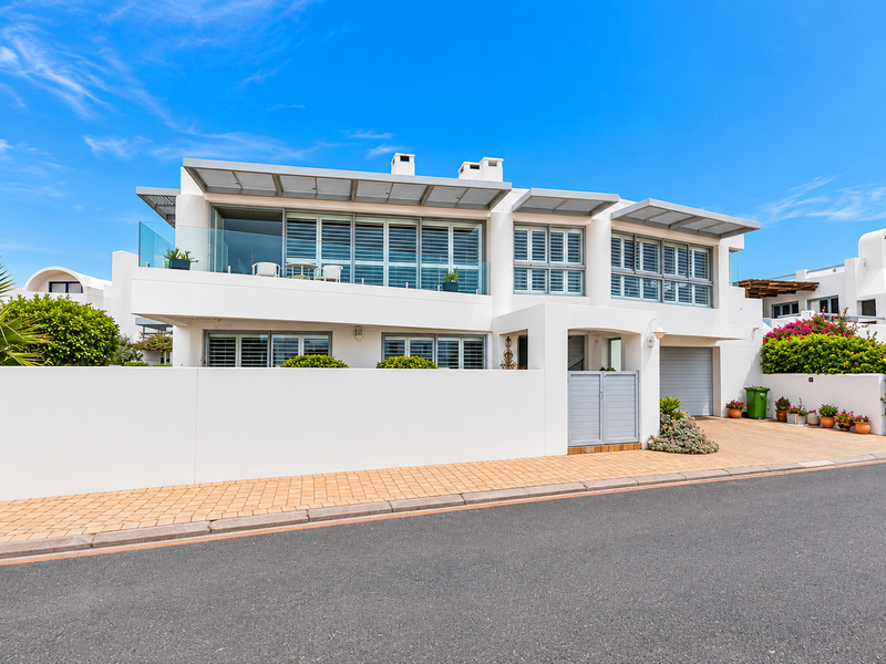 Modern two-story residential facade featuring a paved driveway, integrated garage, and upper-level balcony with glass balustrades and louvre shutters.