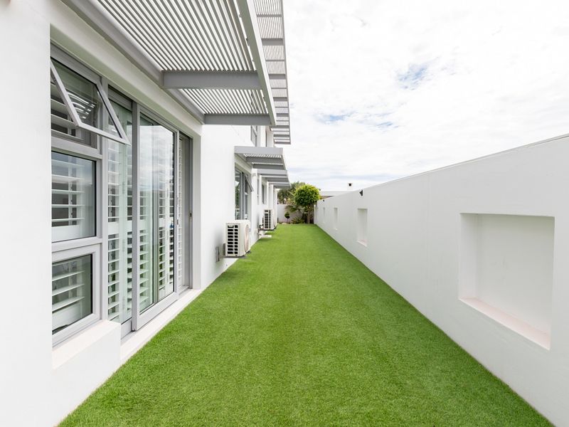 A clean, modern rear exterior space featuring artificial turf, sliding glass doors, and a sleek louvre pergola, enclosed by a high boundary wall.