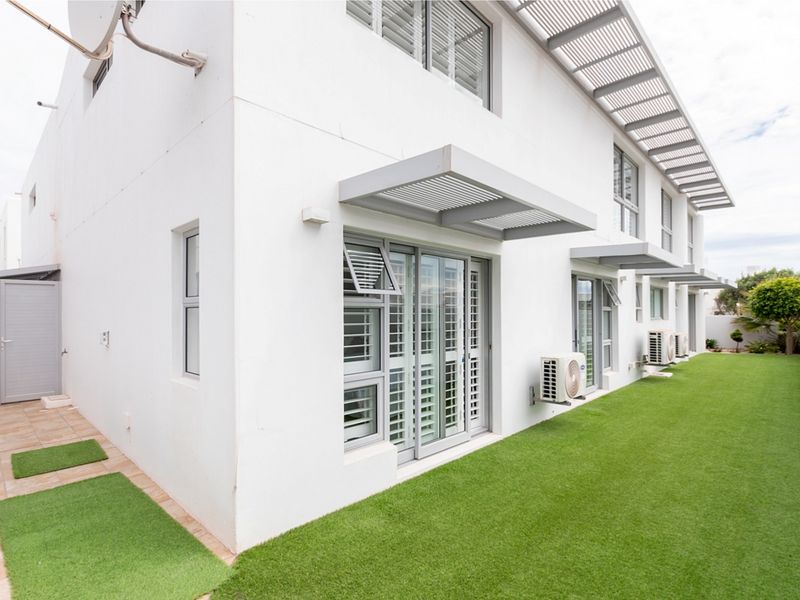Rear elevation of a modern house featuring sliding glass doors, protective louvre awnings, external air conditioning units, and a well-maintained artificial lawn.