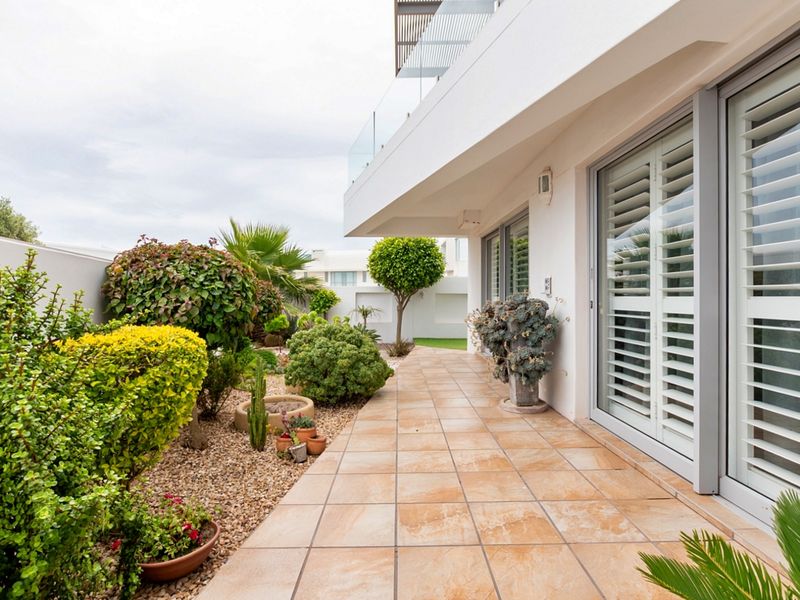 A well-maintained tiled patio area featuring sliding glass doors with plantation shutters, leading to a landscaped garden with a glass balustrade above.