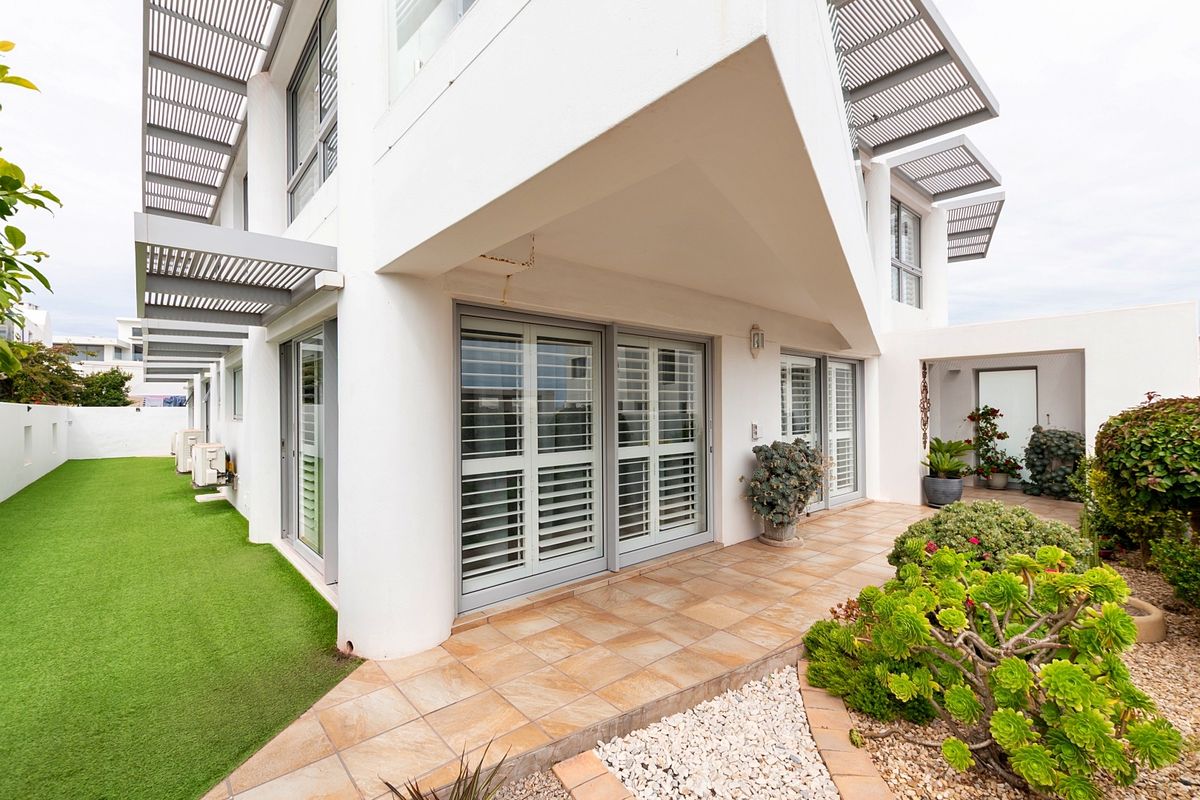 Rear exterior view featuring a tiled patio, sliding doors with shutters, artificial lawn, and modern architectural metal pergola structures.