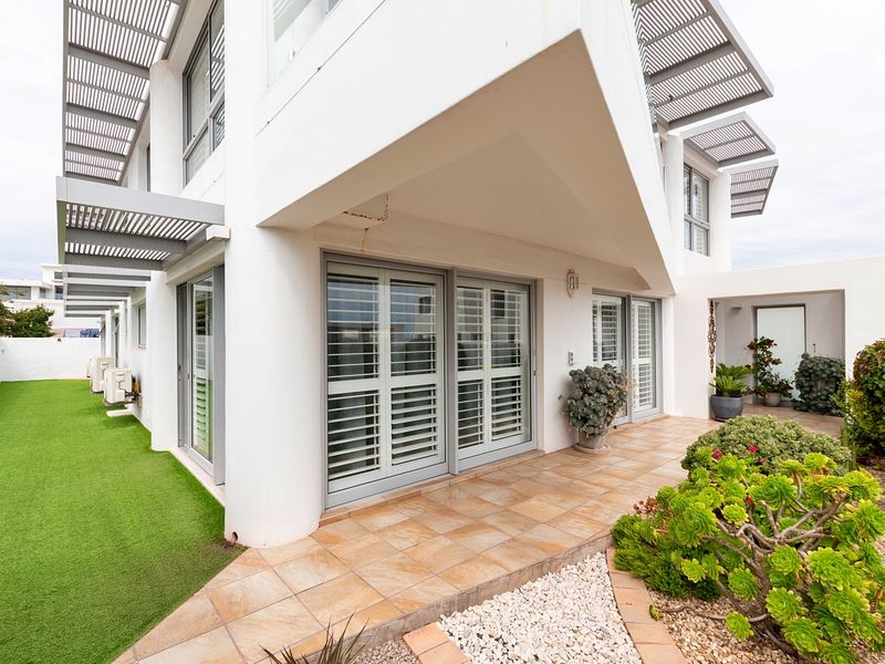 Rear exterior view featuring a tiled patio, sliding doors with shutters, artificial lawn, and modern architectural metal pergola structures.