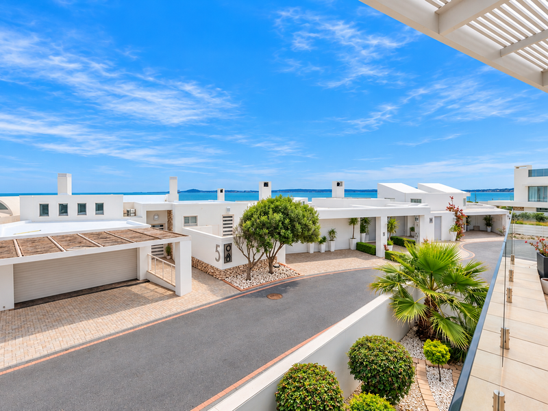 Elevated view overlooking a modern residential complex with paved driveways, garages, and a scenic coastal backdrop.