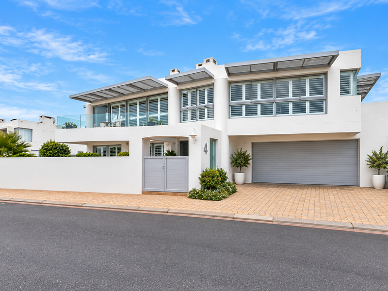Modern two-story residential facade featuring a double garage, paved driveway, and upper-level balcony with glass balustrades and adjustable louvre shutters.