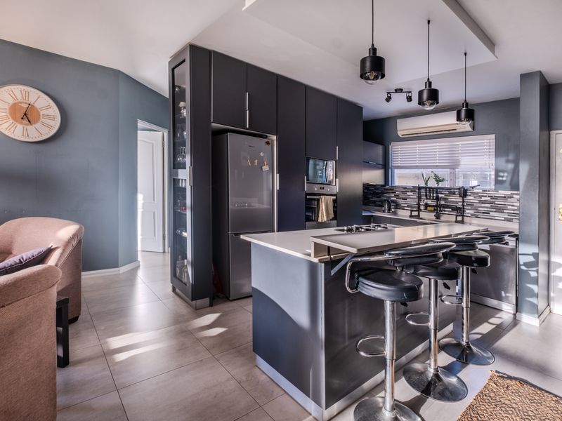 Modern open-plan kitchen featuring dark cabinetry, stone countertops, a central island with breakfast bar, and integrated appliances, seamlessly flowing into the living area.