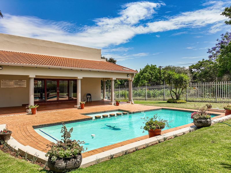 A well-maintained outdoor pool area featuring a tiled patio, covered veranda, and landscaped garden surroundings.