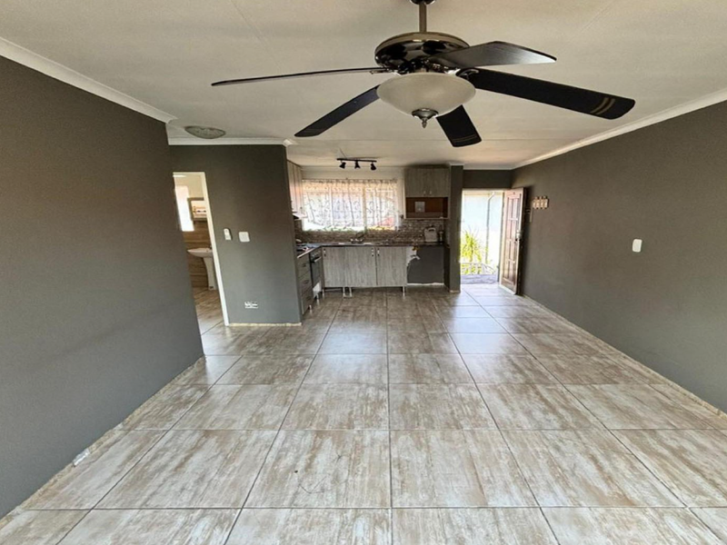 An open-plan living and kitchen area featuring tiled flooring, a ceiling fan, and built-in kitchen cabinetry in need of cosmetic attention.