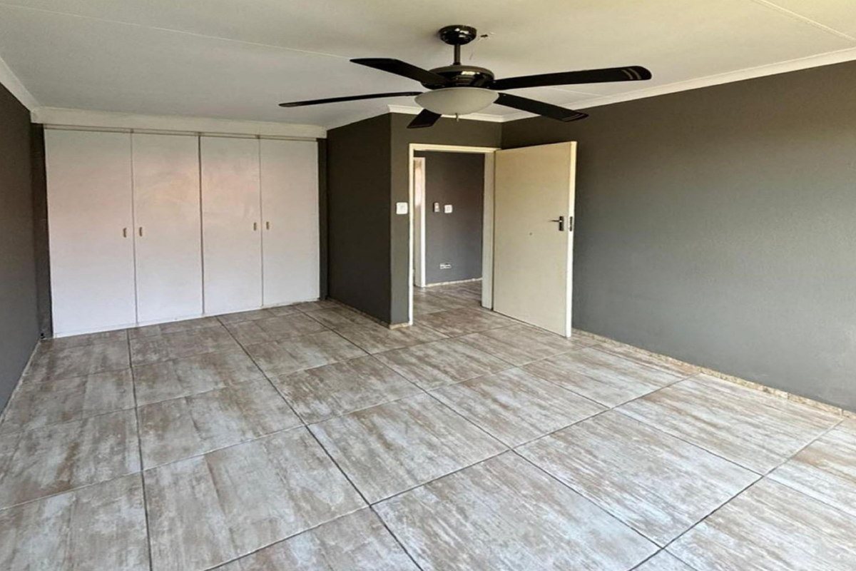 A spacious, empty bedroom featuring neutral grey walls, large format tiled flooring, built-in cupboards, and a central ceiling fan.