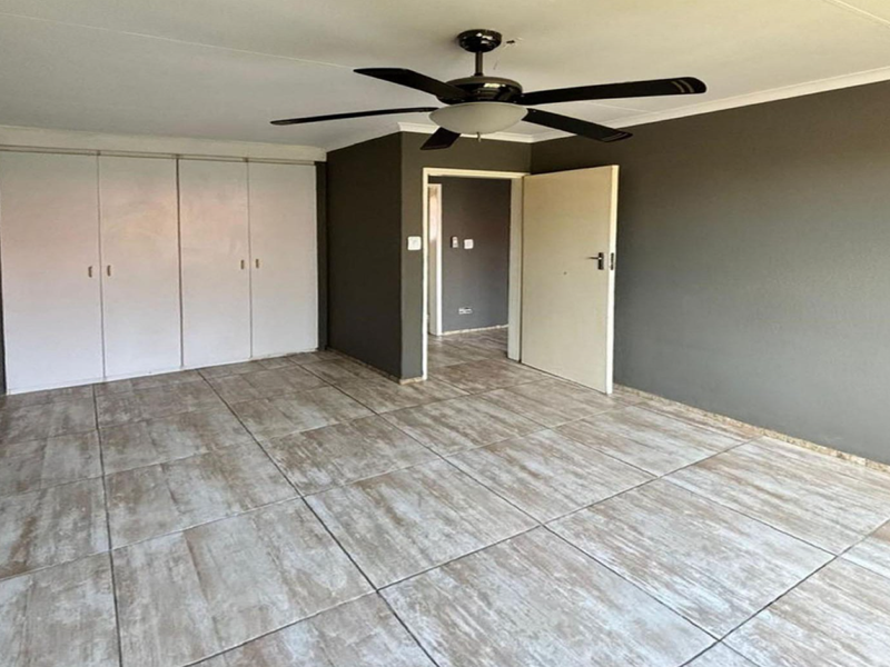A spacious, empty bedroom featuring neutral grey walls, large format tiled flooring, built-in cupboards, and a central ceiling fan.