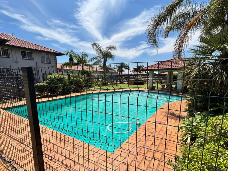 A well-maintained communal swimming pool area featuring brick paving, secure metal fencing, and a covered gazebo for outdoor relaxation.