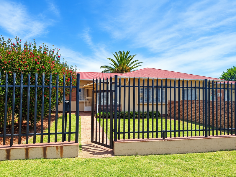 Front view of a townhouse featuring a secure metal fence, brick boundary wall, paved walkway, and a red corrugated metal roof.
