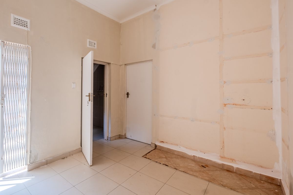 An empty bedroom featuring tiled flooring, security gate, and internal doors, currently requiring wall finishing and floor repairs to restore the space.