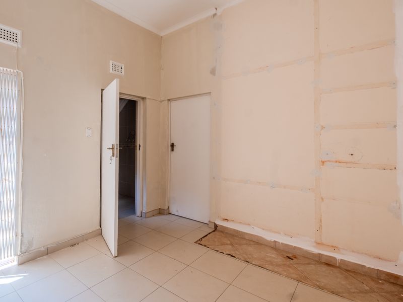 An empty bedroom featuring tiled flooring, security gate, and internal doors, currently requiring wall finishing and floor repairs to restore the space.