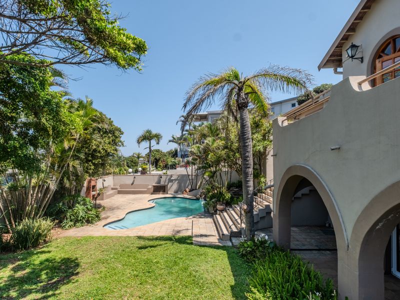 A spacious outdoor pool area featuring a swimming pool, paved patio, and built-in braai, set within a landscaped garden with architectural arched features.