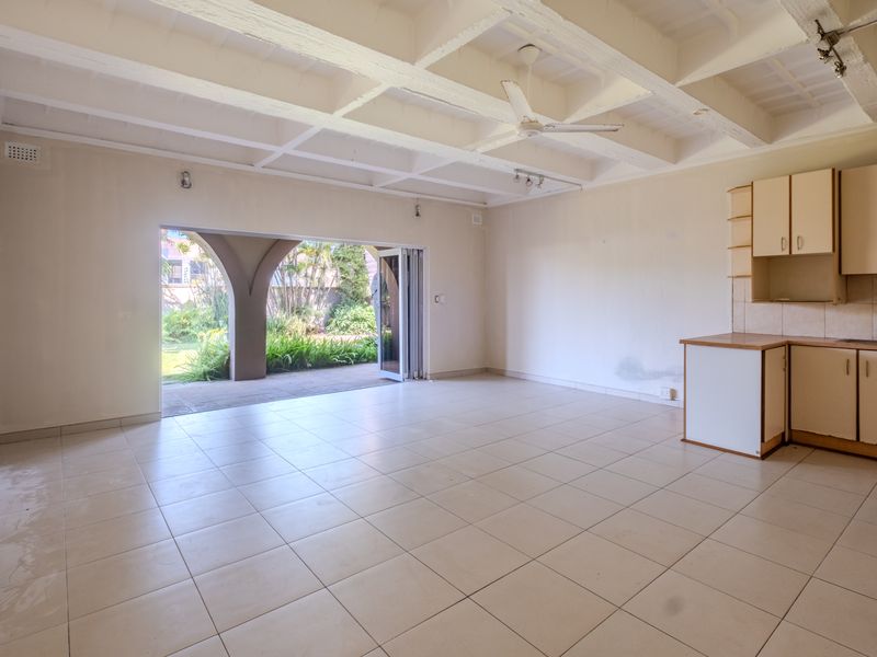 Spacious open-plan living area featuring exposed beam ceilings, tiled floors, and built-in kitchen cabinetry, with seamless access to a covered patio and garden.