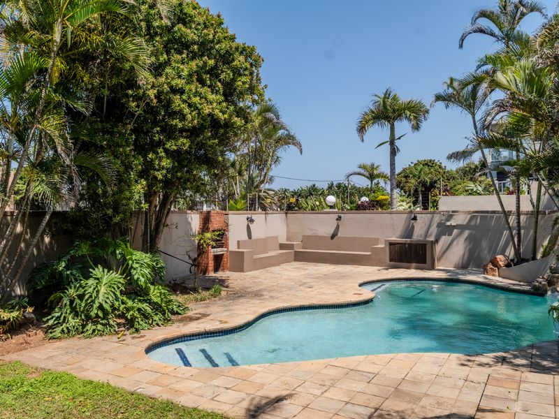Outdoor pool area featuring a swimming pool, paved deck, built-in brick braai, and integrated concrete seating, surrounded by a private boundary wall and garden.