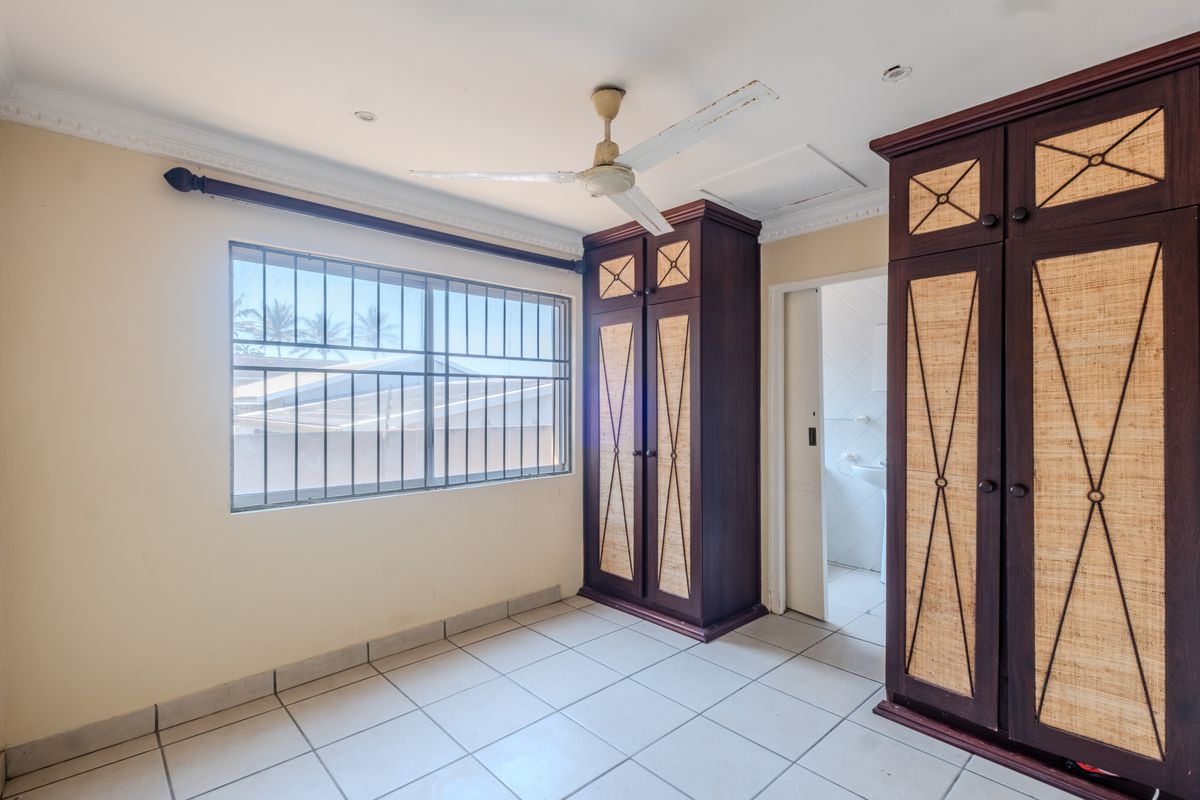 A vacant bedroom featuring tiled flooring, built-in wooden cupboards, a ceiling fan, and security bars on the window.