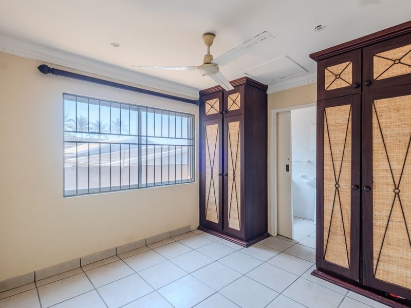 A vacant bedroom featuring tiled flooring, built-in wooden cupboards, a ceiling fan, and security bars on the window.