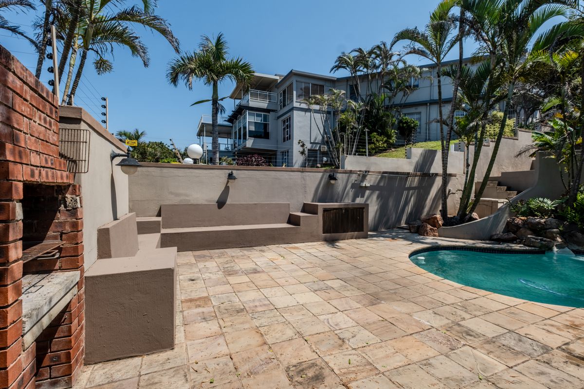 Outdoor pool area featuring a paved patio, built-in braai, and integrated seating, set against a retaining wall with established palm trees.