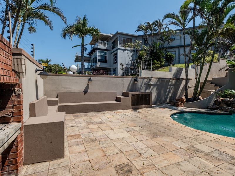 Outdoor pool area featuring a paved patio, built-in braai, and integrated seating, set against a retaining wall with established palm trees.