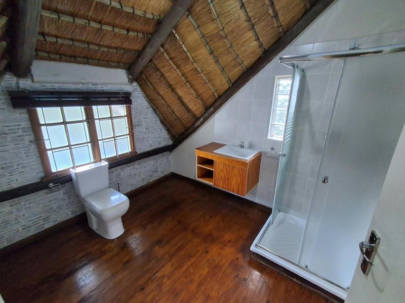 A unique attic-style bathroom featuring a thatched roof, exposed timber beams, timber flooring, a modern shower enclosure, and a wall-mounted vanity unit.