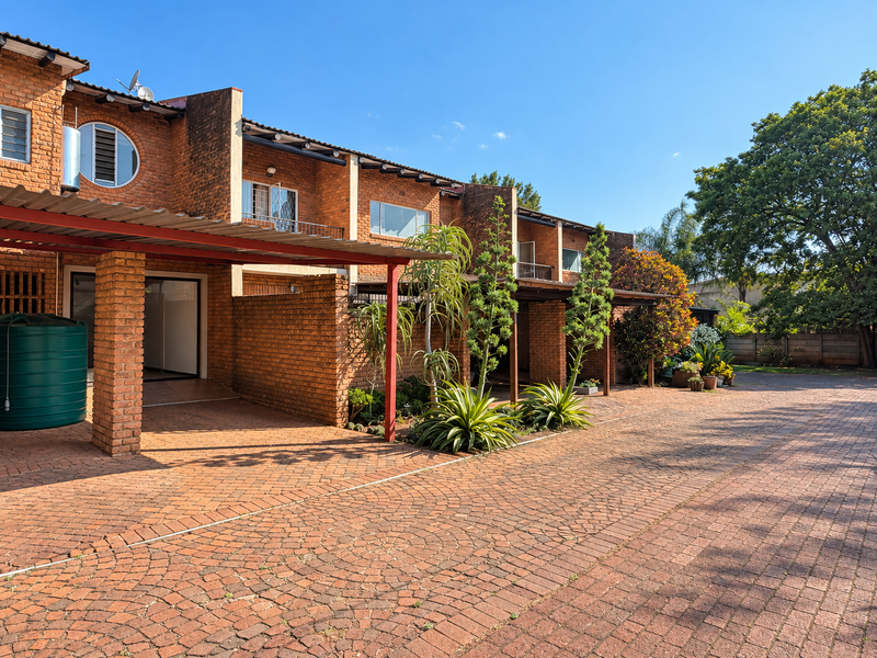 Front exterior view of a brick townhouse complex featuring a paved driveway, covered carport, and established garden landscaping.