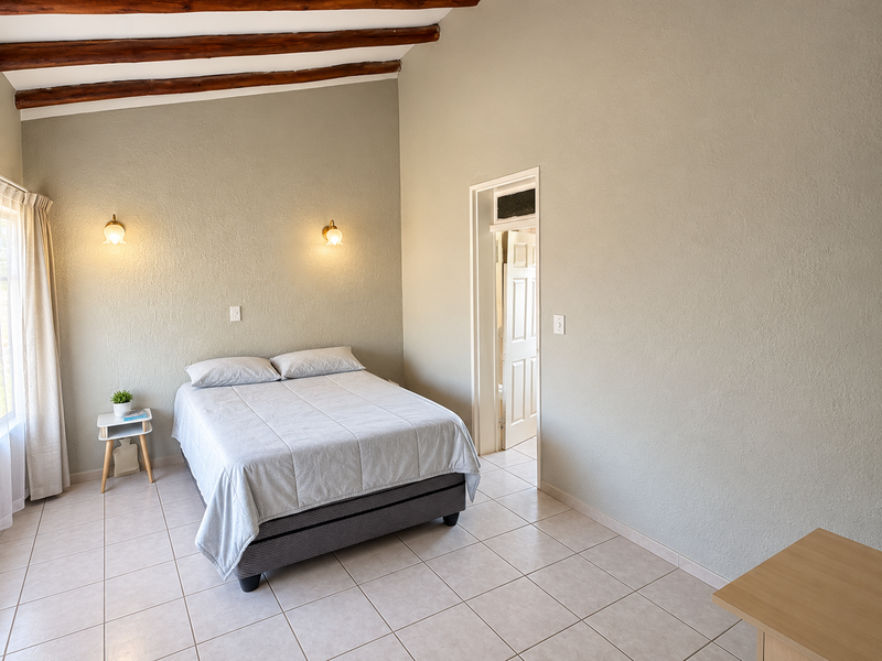 A well-lit bedroom featuring exposed timber ceiling beams, neutral wall finishes, and tiled flooring, with direct access to an adjoining room.