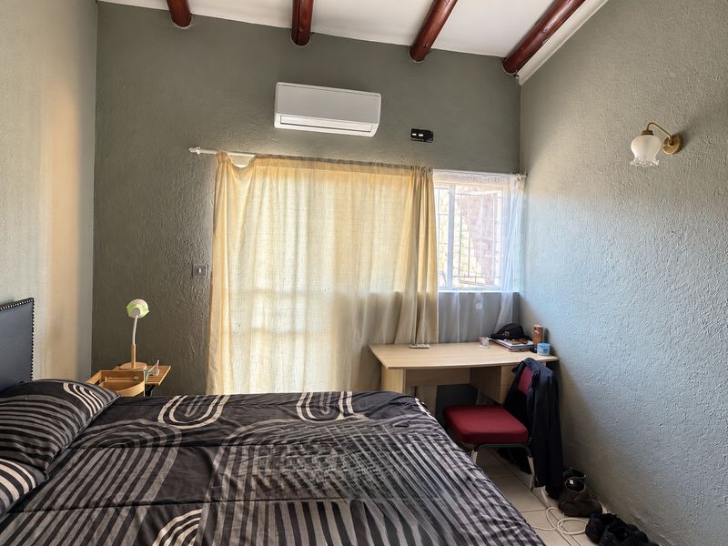 A bedroom featuring exposed timber ceiling beams, a split-system air conditioner, and a window with security bars.