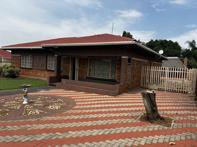 Front view of a single-story brick house featuring a tiled roof, covered porch, and a paved driveway with perimeter fencing.
