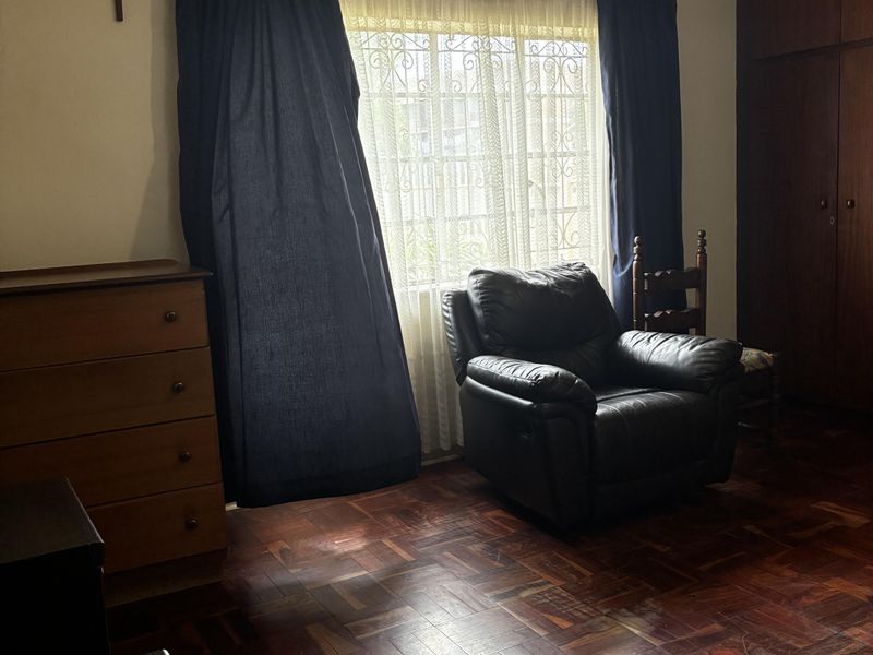 A bedroom featuring classic parquet flooring, built-in wooden cupboards, and a ceiling fan, offering a traditional layout with natural light through a security-gated window.