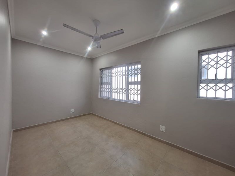 A clean, empty bedroom featuring neutral tiled flooring, recessed downlighting, a ceiling fan, and windows fitted with security gates.