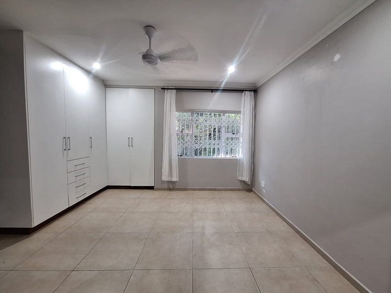 A spacious, empty bedroom featuring neutral tiled flooring, extensive white built-in cupboards, a ceiling fan, and a window fitted with a security gate.